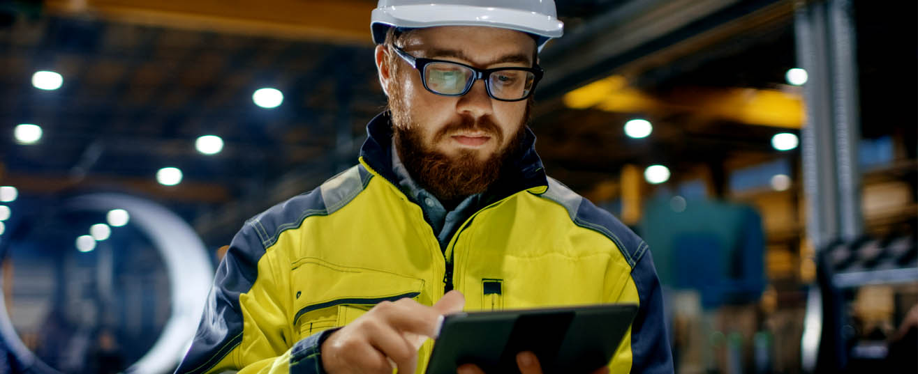 Man in high visibility jacket and hard hat studying a screen in a warehouse.